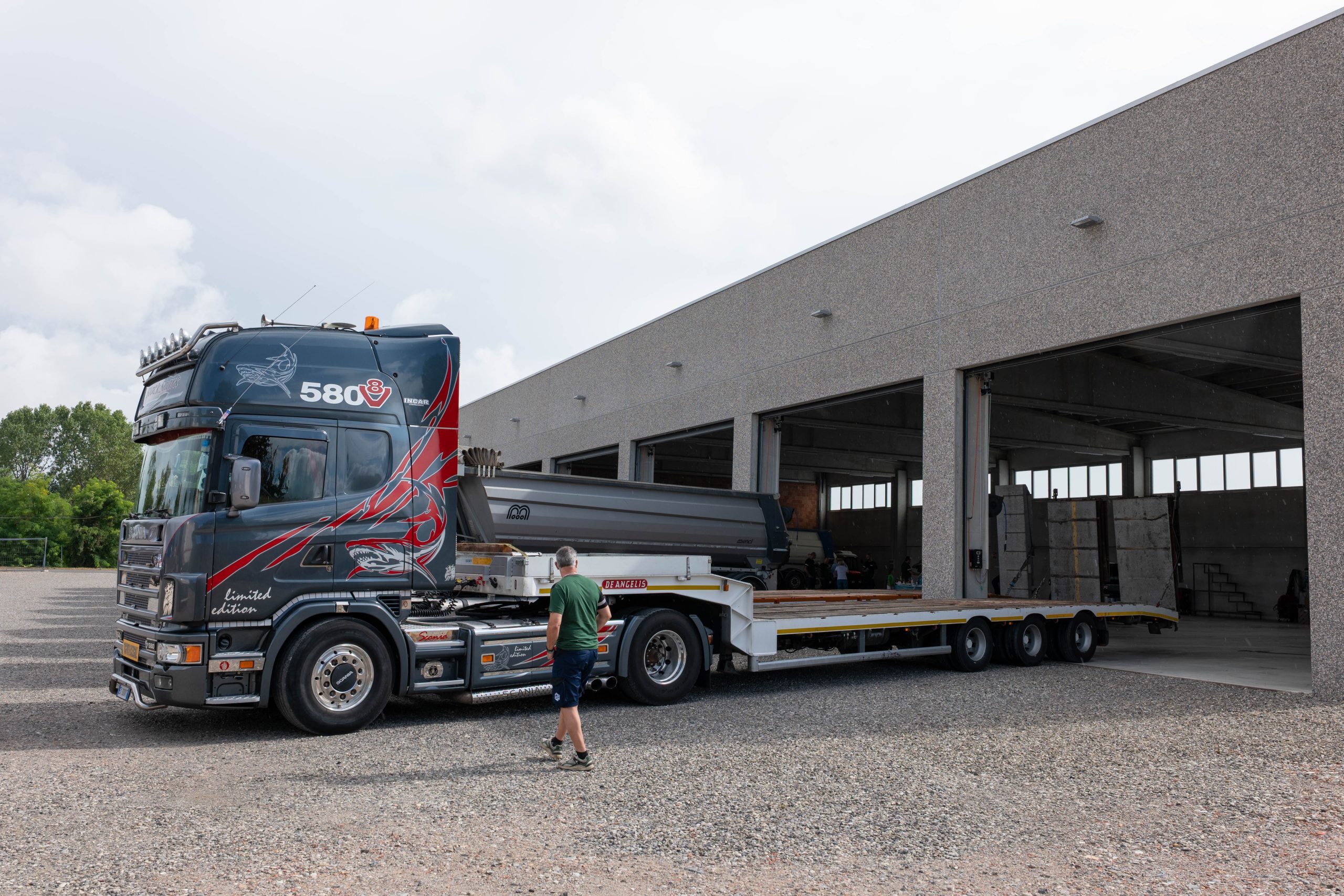 Camion Scania pianale con rampa ribassato parcheggiato davanti a un capannone